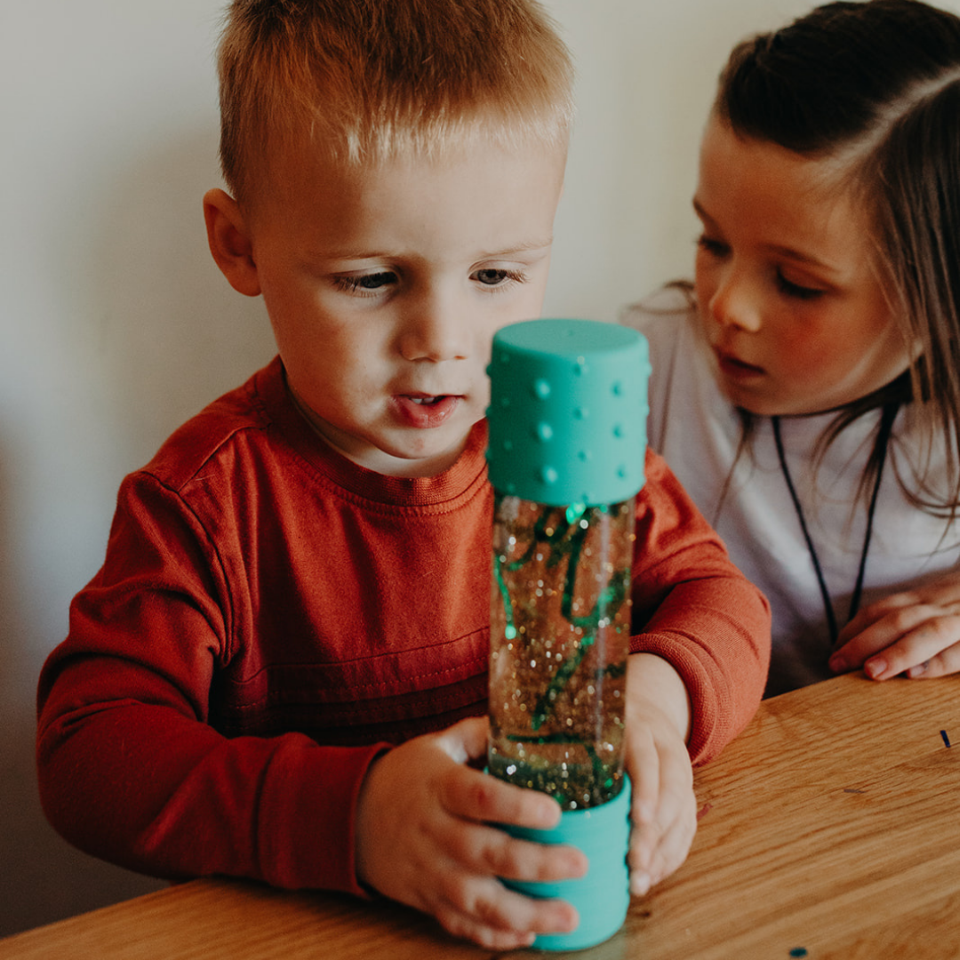 Two children looking at a glittery bottle with a teal top on a wooden table.