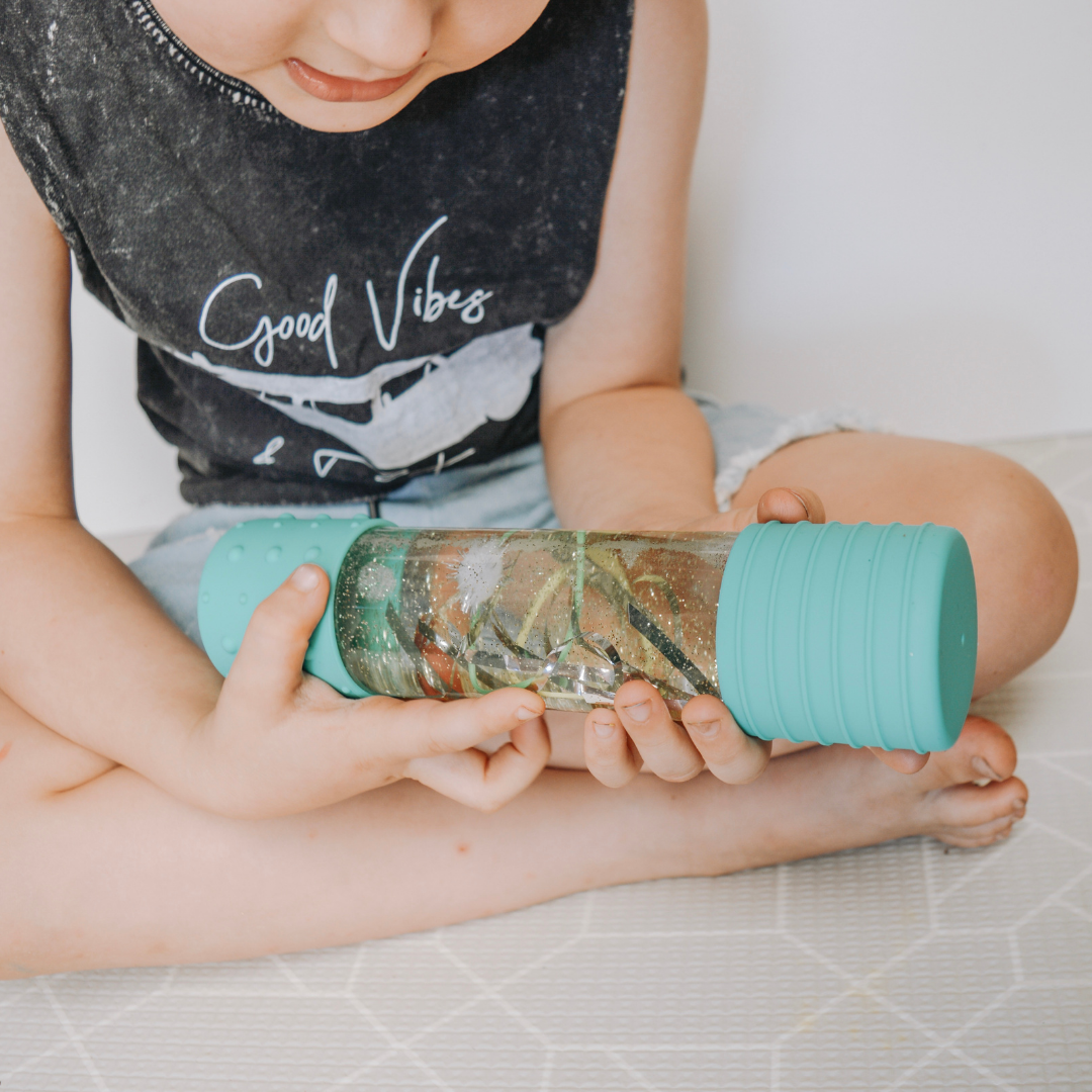 Child holding a teal water bottle with a clear lid on a light-colored floor.