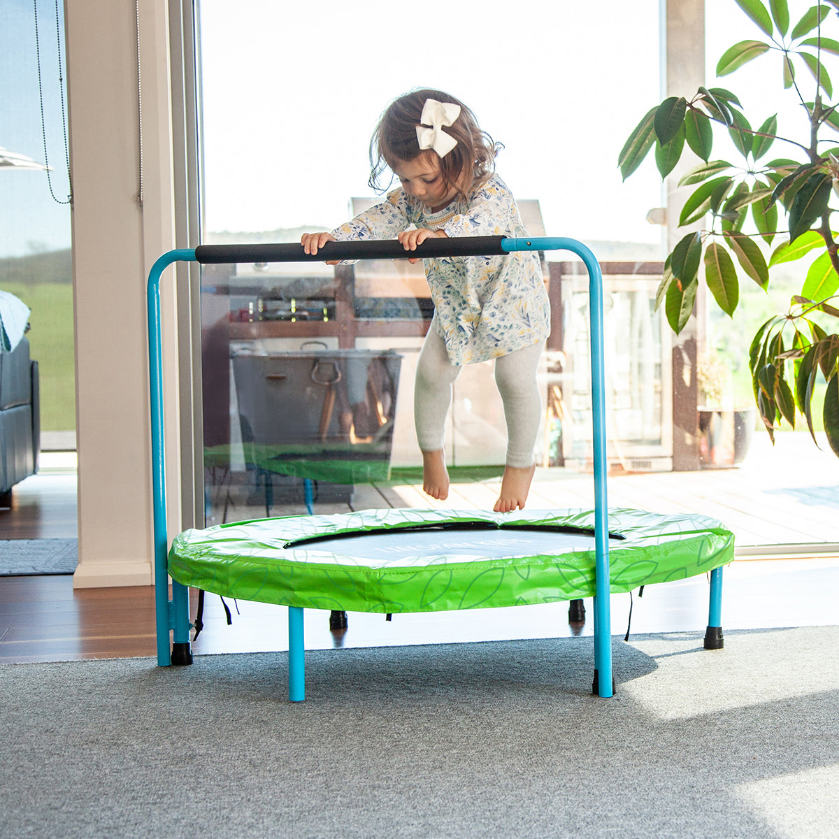 Child on a small green trampoline indoors with a plant and furniture in the background