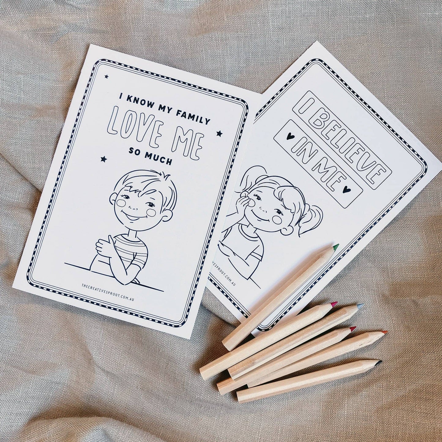 Two coloring books with illustrations and text on a textured fabric background, accompanied by wooden crayons.