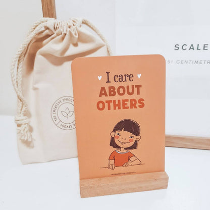 Small book with 'I care ABOUT OTHERS' text and a cartoon character on an orange background, next to a beige drawstring bag.
